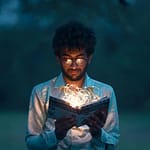 A man with glasses reads an illuminated book outdoors during twilight.
