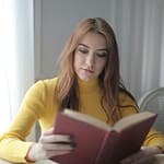 A young woman in a yellow sweater reads a book indoors, enjoying a calm and leisurely moment.