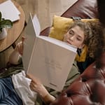 A young woman is lying on a sofa reading a book in a cozy indoor setting.