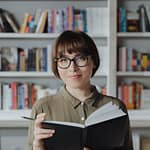 Smiling woman in a library holding a book, surrounded by bookshelves.