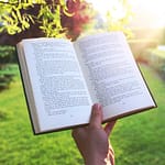 Open book held by hand in a bright green garden under sunlight, evoking relaxation and leisure.