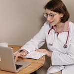 Female doctor using a laptop while sitting at a desk with notes.
