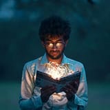 A man with glasses reads an illuminated book outdoors during twilight.