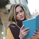 A young woman enjoys reading a book while listening to music on a city street.
