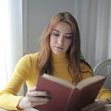 A young woman in a yellow sweater reads a book indoors, enjoying a calm and leisurely moment.