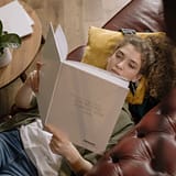 A young woman is lying on a sofa reading a book in a cozy indoor setting.
