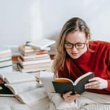 A woman in a red sweater reading a book on the floor with stacks of books around her indoors.
