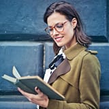 Smiling woman enjoying a book while standing against a textured blue wall.