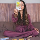 A woman sitting indoors in a purple dress holding up a tarot card, suggesting a spiritual reading.