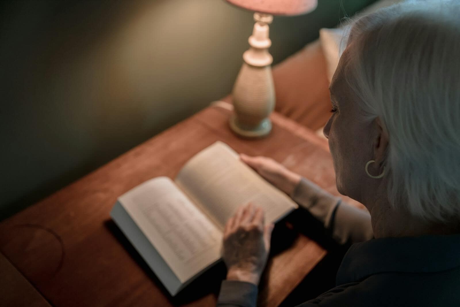 Elderly woman enjoying a quiet reading moment beside a table lamp indoors.
