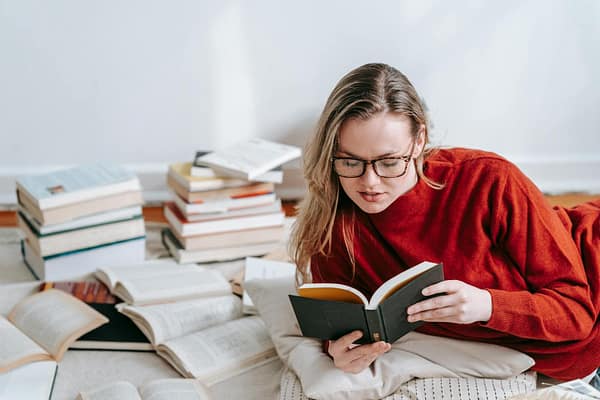 A woman in a red sweater reading a book on the floor with stacks of books around her indoors.