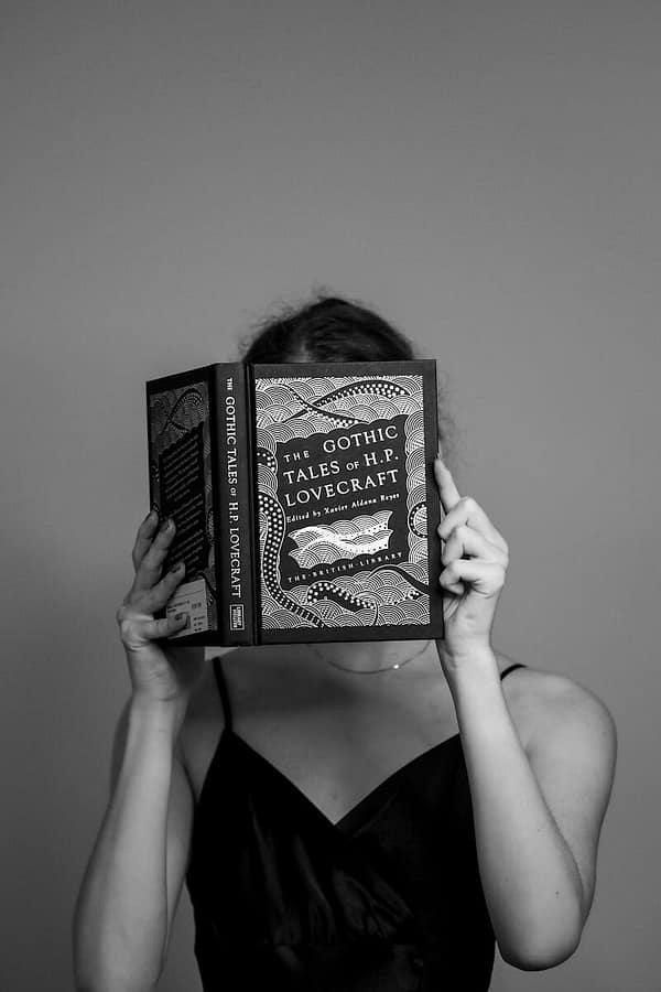 Black and white portrait of a woman reading 'The Gothic Tales of H.P. Lovecraft' in a studio setting.