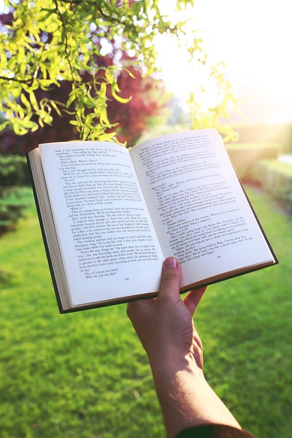 Open book held by hand in a bright green garden under sunlight, evoking relaxation and leisure.