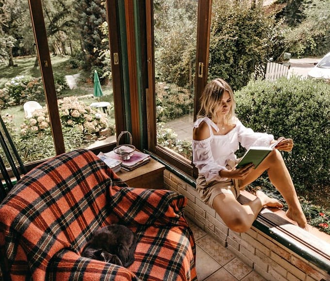 Woman enjoying a relaxing afternoon reading by a window with a garden view.