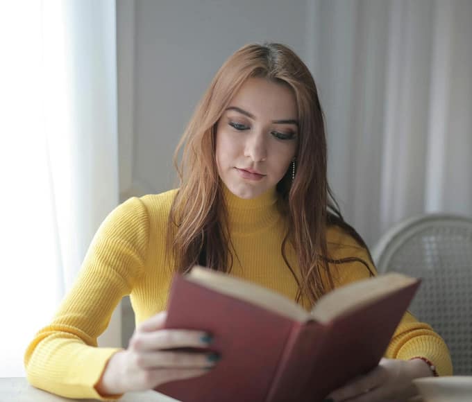 A young woman in a yellow sweater reads a book indoors, enjoying a calm and leisurely moment.