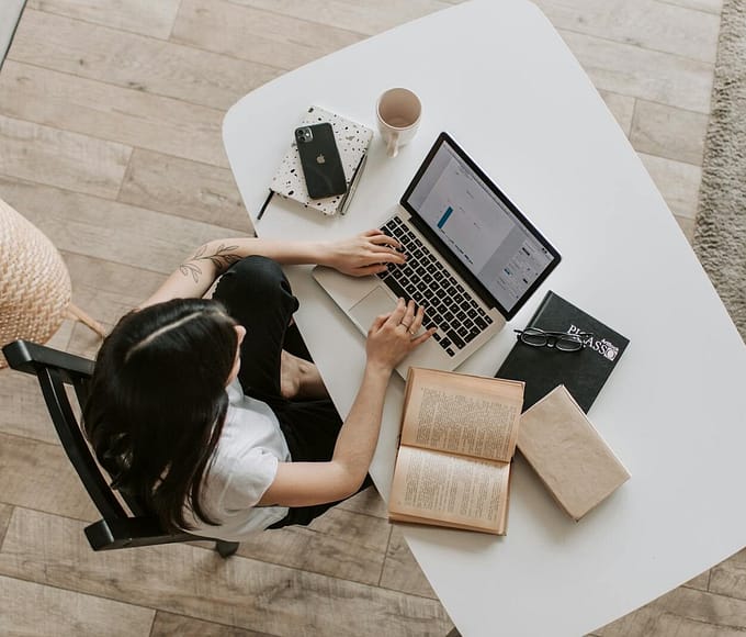 Overhead view of a woman using a laptop at a home desk, surrounded by books, a phone, and a cup.