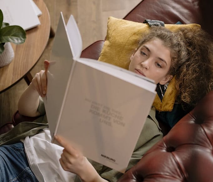 A young woman is lying on a sofa reading a book in a cozy indoor setting.