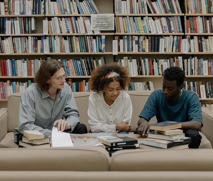 Three students engaged in group study session surrounded by bookshelves.