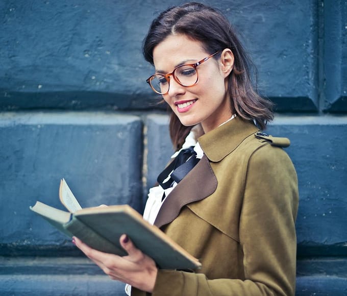 Smiling woman enjoying a book while standing against a textured blue wall.