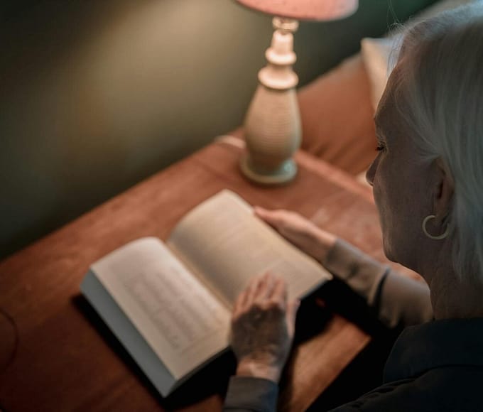 Elderly woman enjoying a quiet reading moment beside a table lamp indoors.