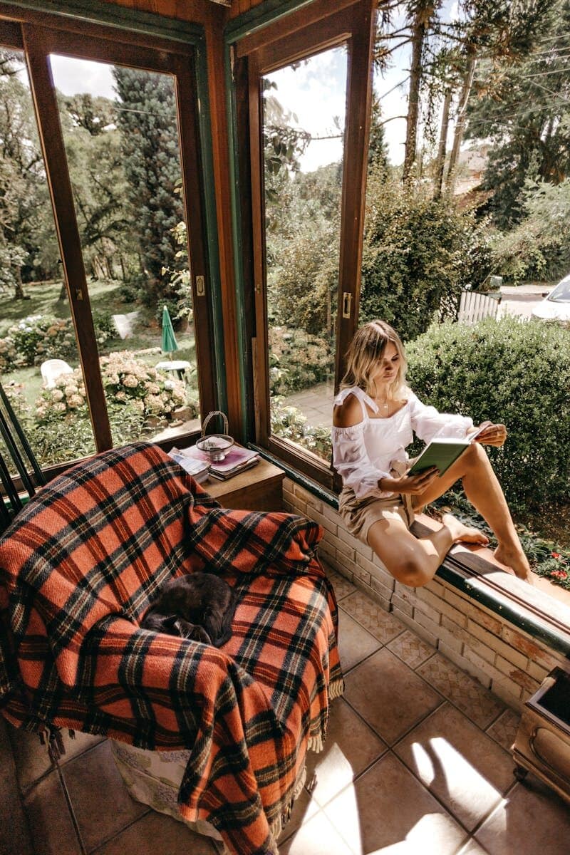 Woman enjoying a relaxing afternoon reading by a window with a garden view.
