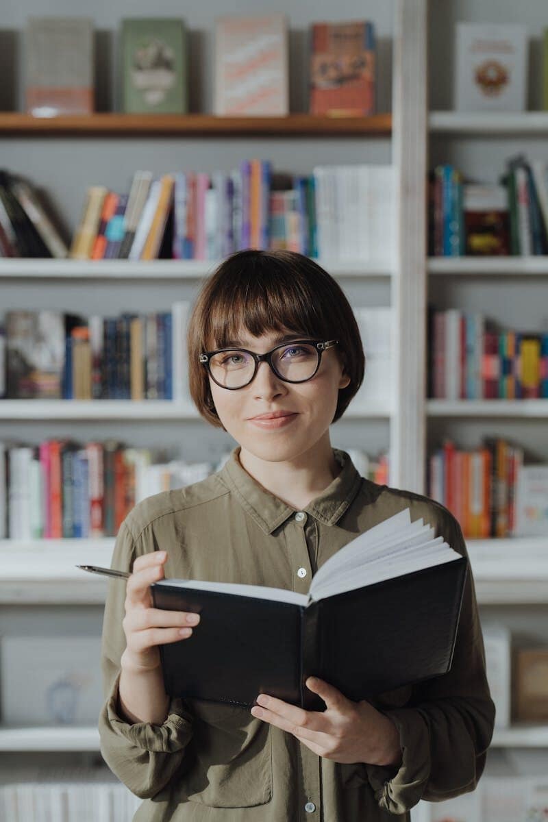 Smiling woman in a library holding a book, surrounded by bookshelves.