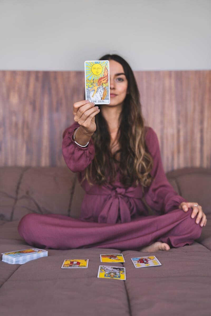 A woman sitting indoors in a purple dress holding up a tarot card, suggesting a spiritual reading.