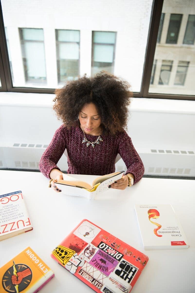 A woman engaged in reading a book at a white desk, surrounded by colorful books, in a modern office space.