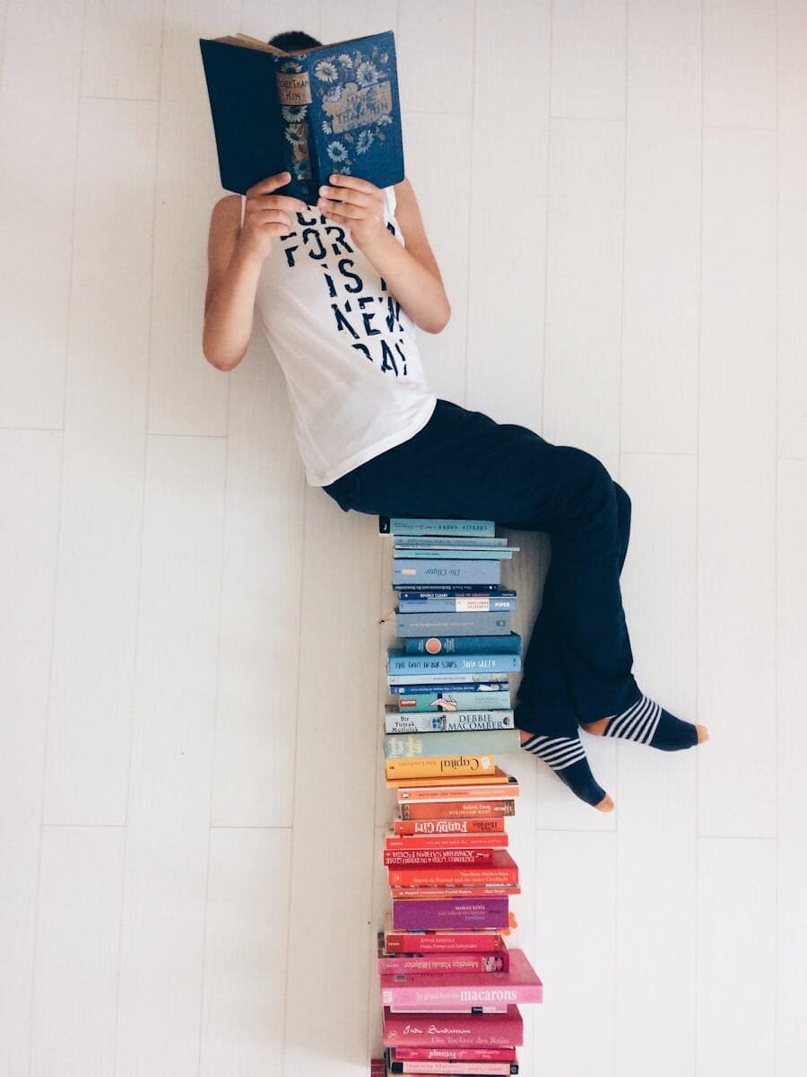 Person casually reading a book while sitting atop a colorful stack of books indoors.