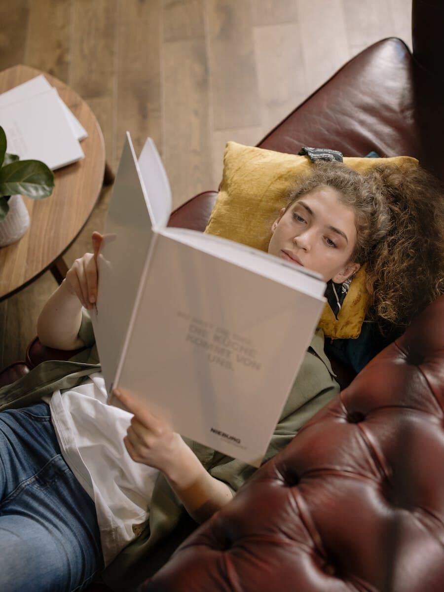 A young woman is lying on a sofa reading a book in a cozy indoor setting.
